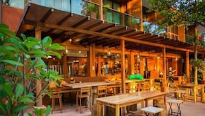 Un área de comedor al aire libre con mesas y sillas de madera, rodeada de vegetación y una zona de estar cubierta.