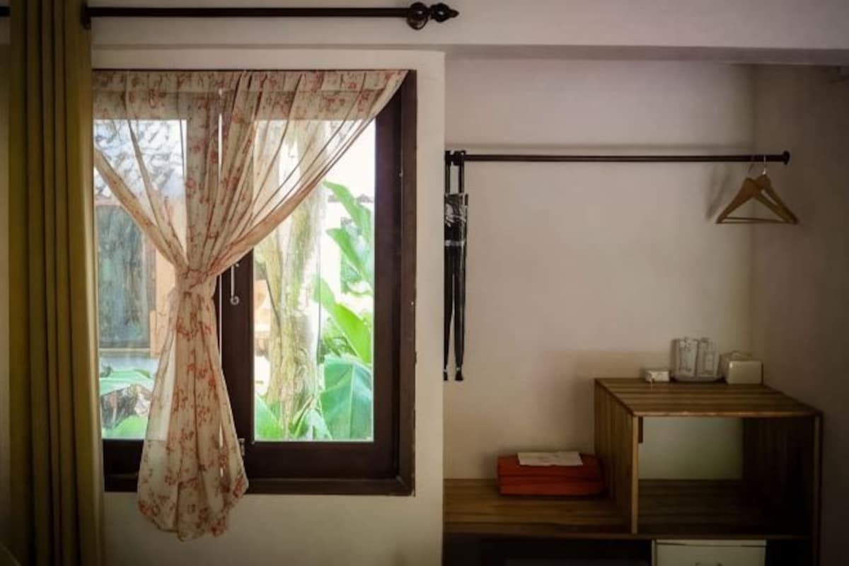 A room with a window, a curtain, a wooden shelf with a red box, and a hanging light fixture.