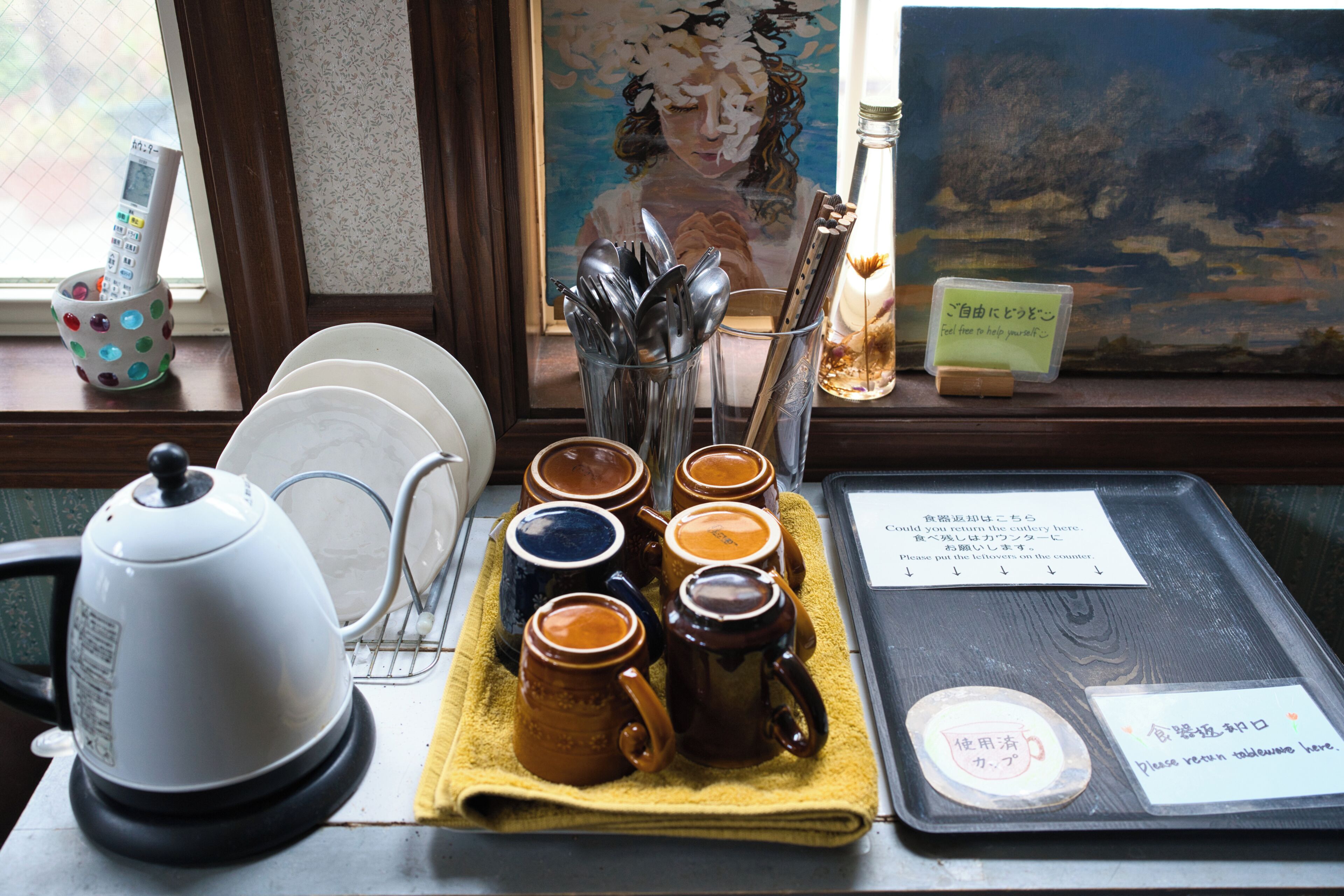 Un plateau avec des tasses à café, une théière et un menu, posé sur une table près d’une fenêtre ornée d’un tableau.