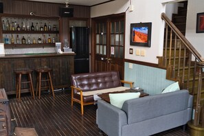 A bar area with shelves of bottles, a refrigerator, wooden stools, a brown leather couch, a gray sofa, and a staircase with wooden railings. - Iza Kamakura Guest House & Bar - Hostel (Kamakura)