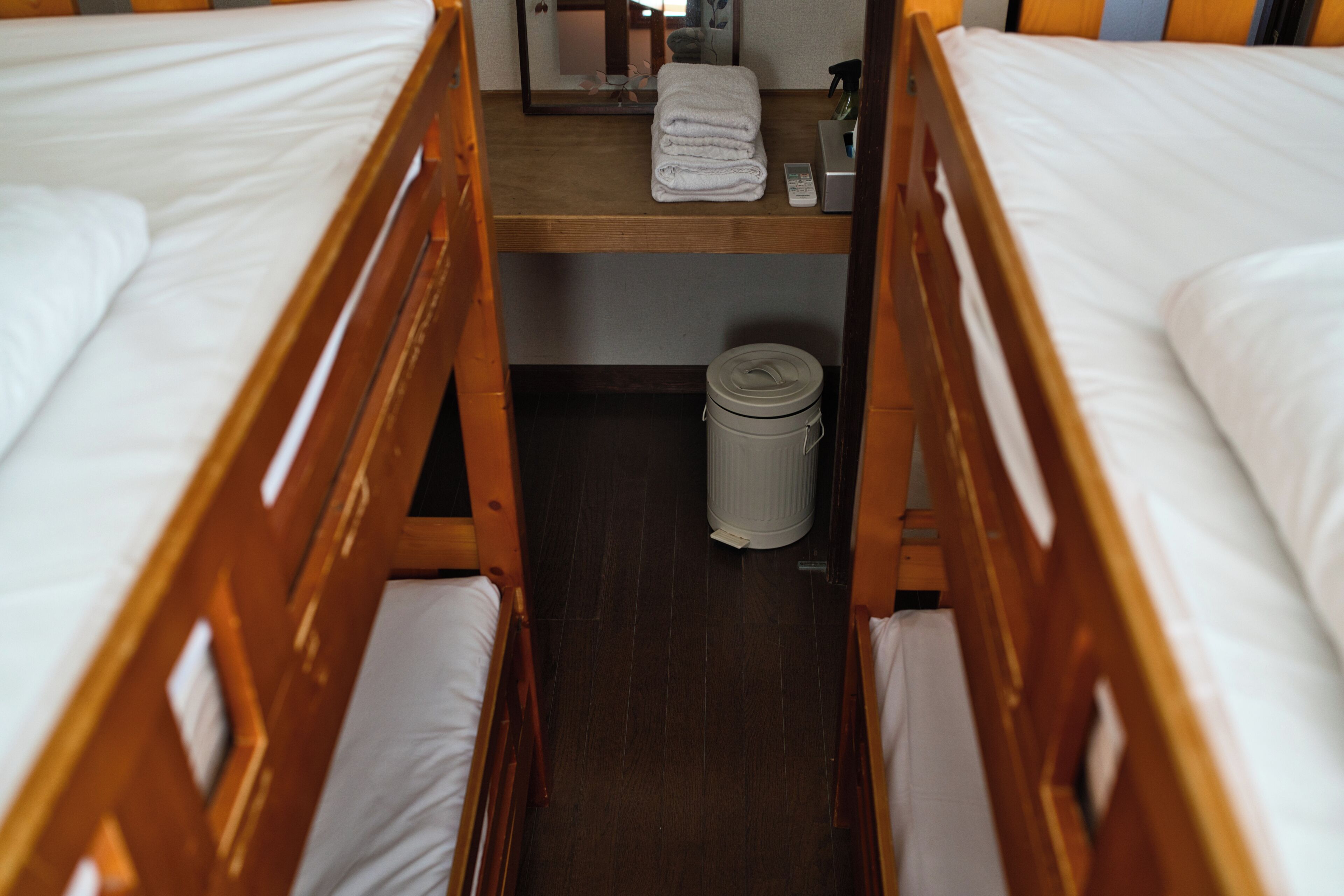 A narrow room with two wooden bunk beds, a small shelf with a folded towel and a remote control, and a trash can.