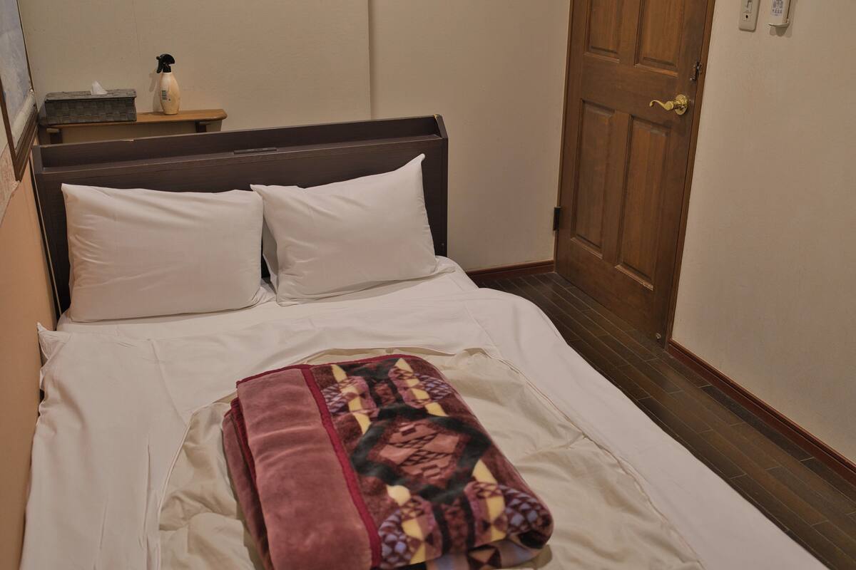 Una cama bien hecha con sábanas blancas, cabecera de madera y una manta decorativa. Se observa una mesita de noche con una botella y pañuelos, y una puerta de madera con pomo de latón.
