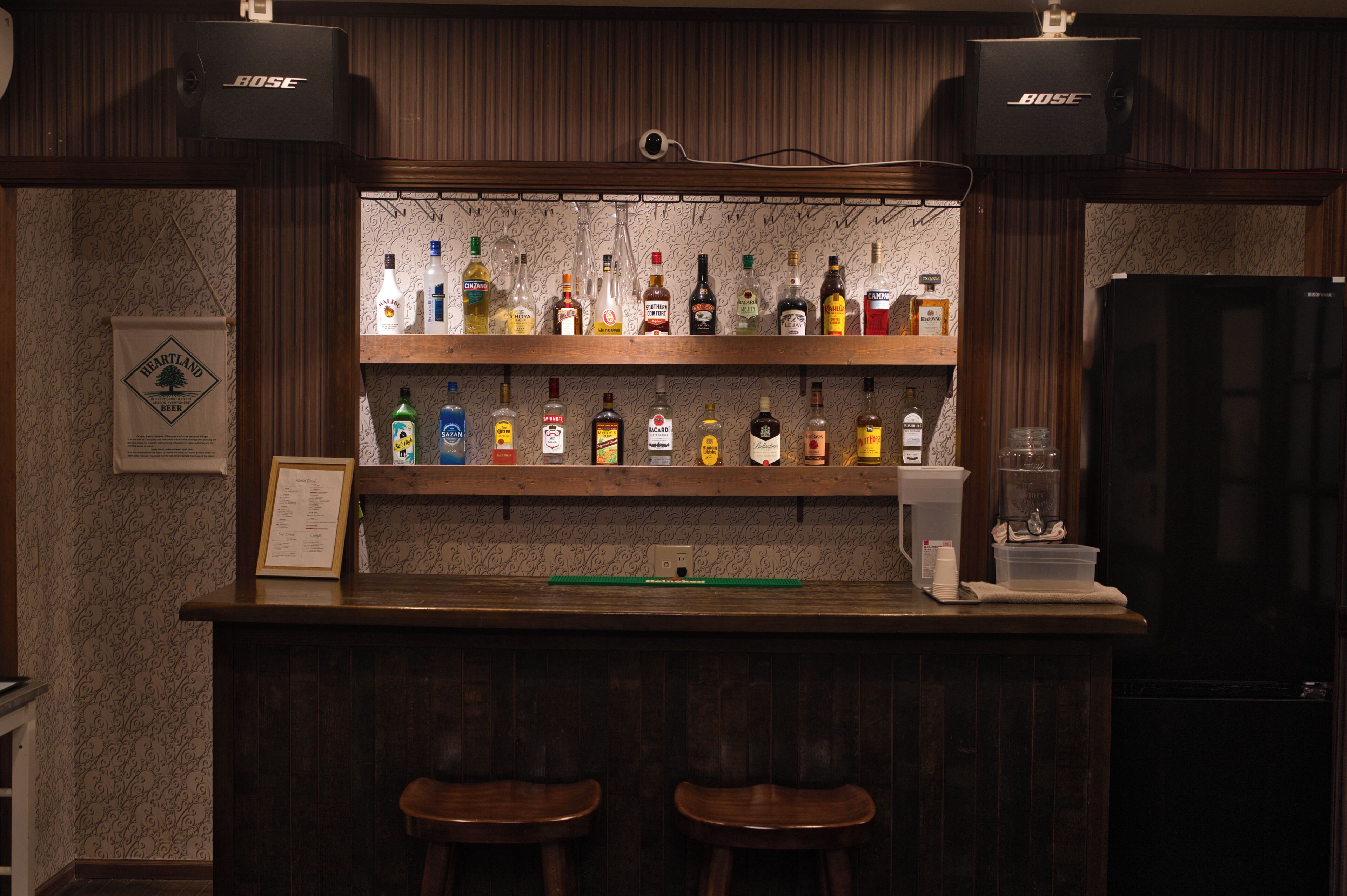 A bar area with wooden shelves stocked with various liquor bottles, two stools, and a refrigerator.