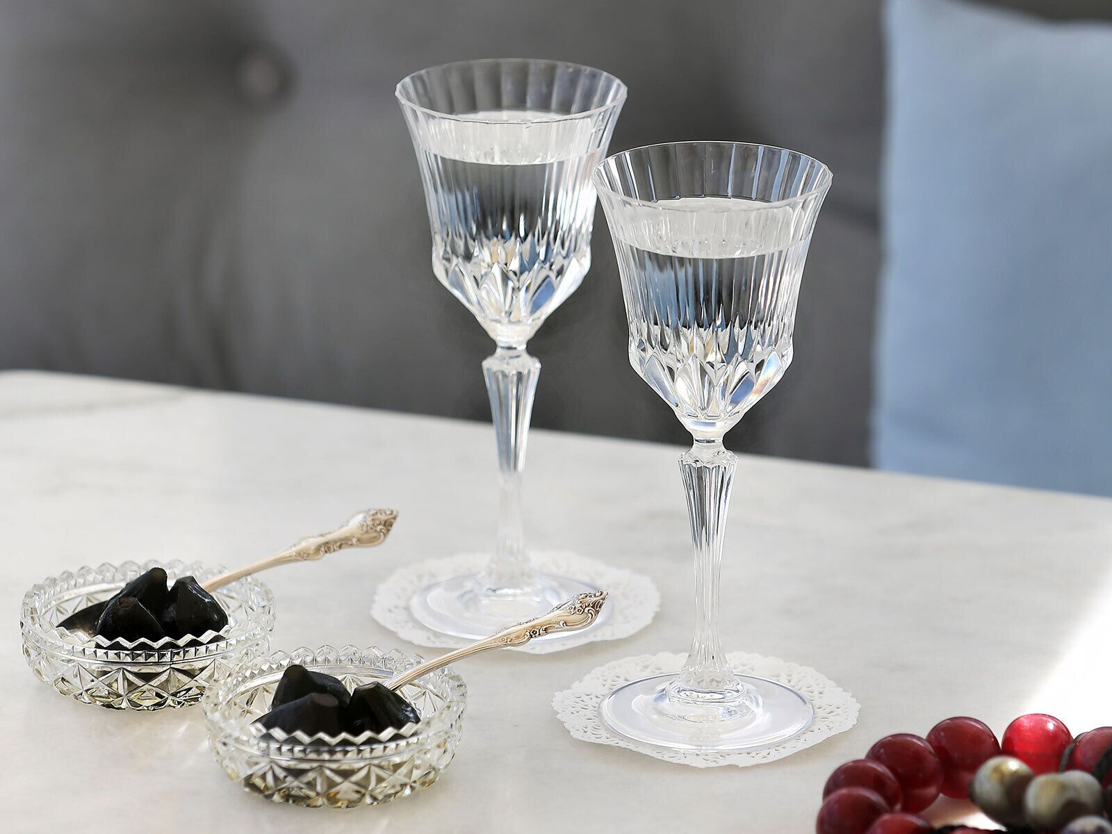 Two crystal glasses on a table with a lace doily, accompanied by a silver spoon and a small bowl with black objects.