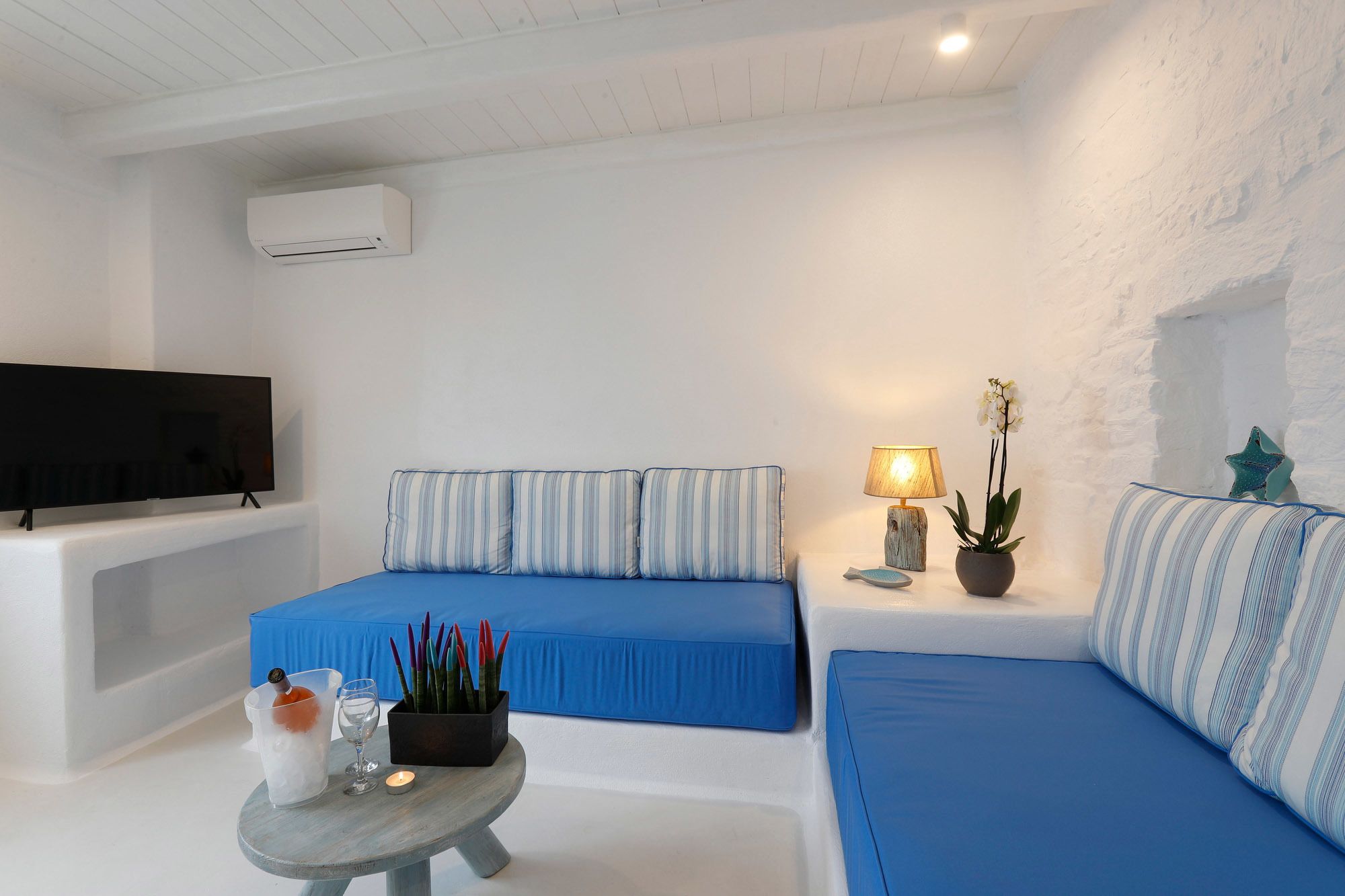 Una sala de estar moderna con un televisor de pantalla plana, dos sofás azules, una mesita redonda con una vela y una planta en maceta.