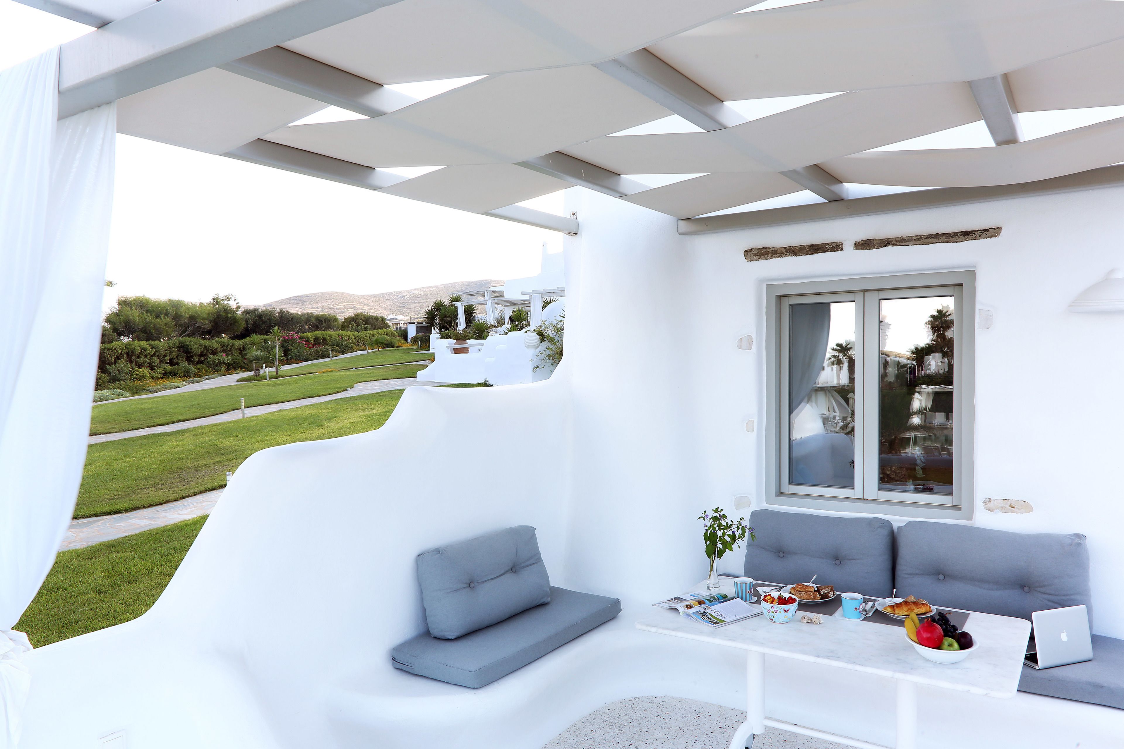 Un área de estar moderna al aire libre con una estructura blanca, cojines grises, una mesa con comida y una computadora portátil, y vista a un paisaje verde.