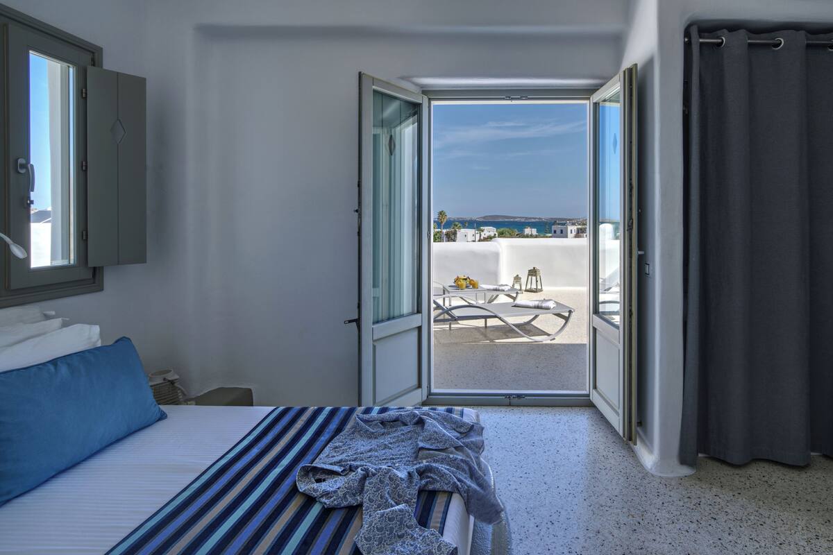 A hotel room with a bed, a window, and an open door leading to a balcony with a view of the sea.