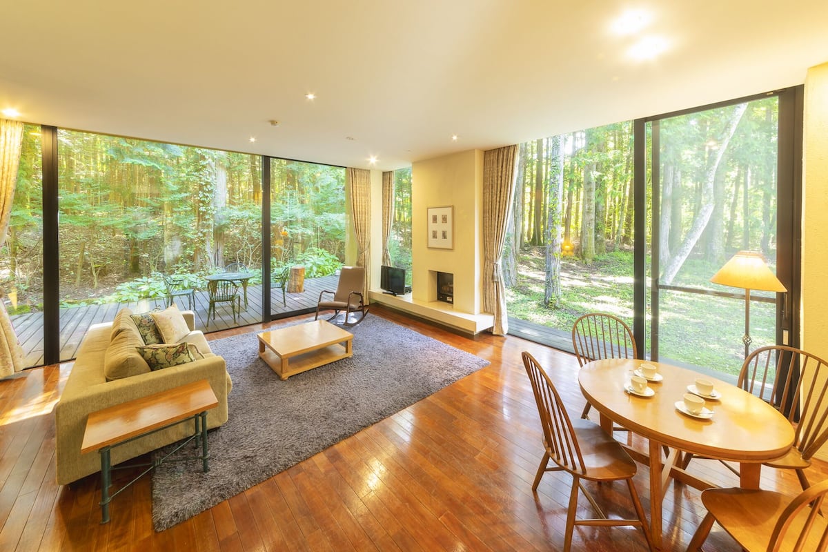 A spacious living room with a large window overlooking a forest, a cozy sofa, a coffee table, and a dining area with chairs.