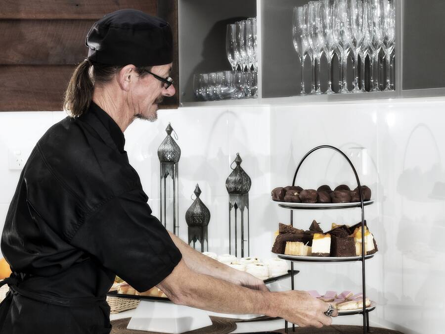 Une personne présente des desserts sur un présentoir à trois niveaux dans une cuisine dont le mur est recouvert de carreaux blancs et orné de supports à bougies en métal décoratif.