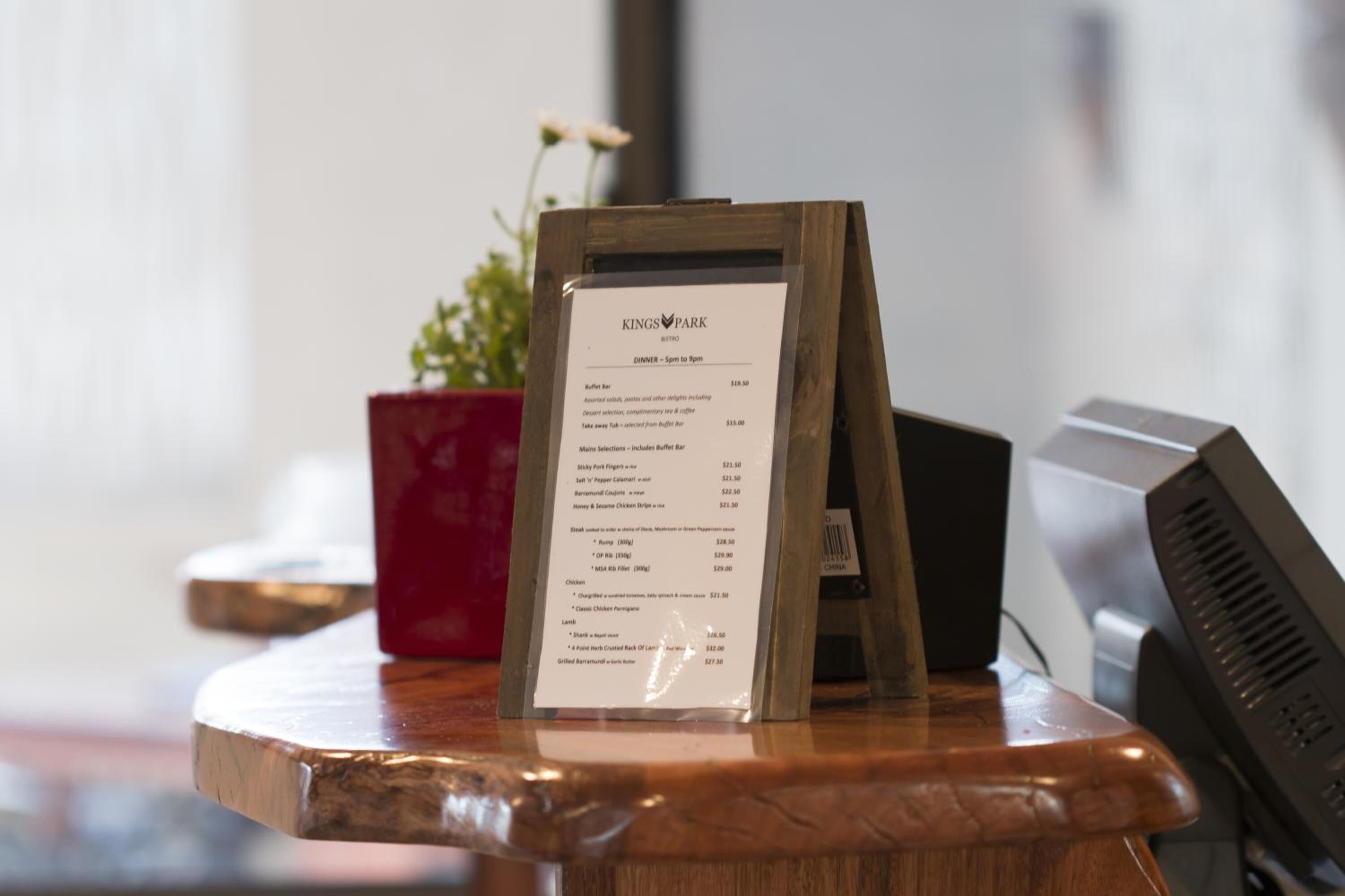 A menu board at a restaurant with a wooden stand, a red pot with flowers, and a computer monitor in the background.