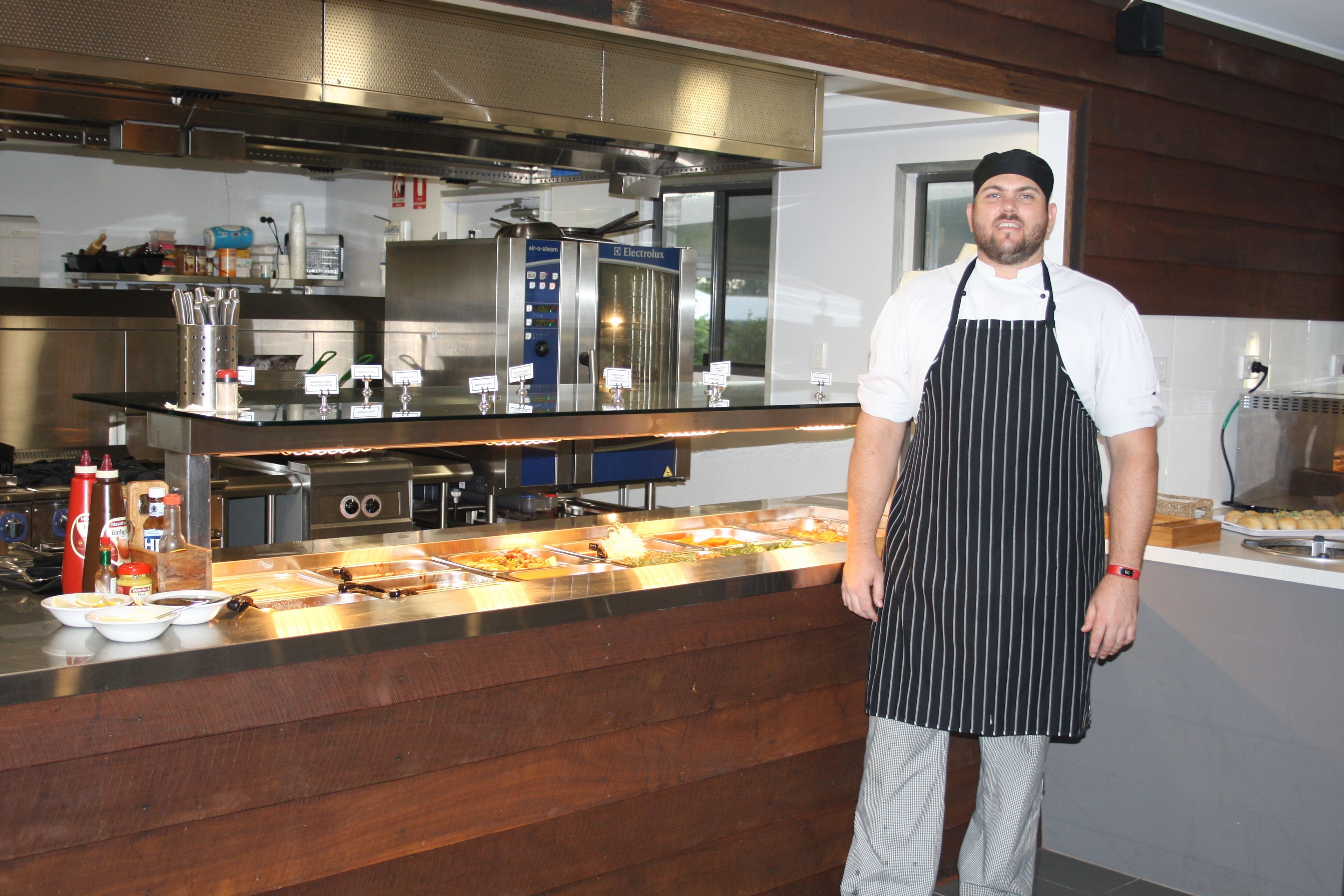 A chef standing in a kitchen with stainless steel appliances, a buffet counter, and wooden paneling.