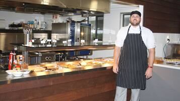 A chef standing in a kitchen with stainless steel appliances, a buffet counter, and wooden paneling.