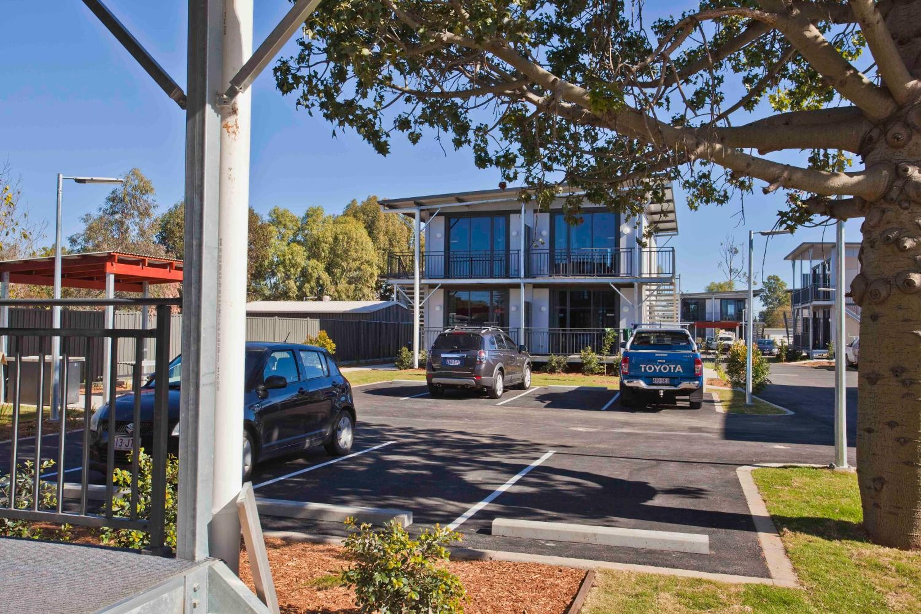 Ein Parkplatz mit Autos, ein Gebäude mit Balkonen und ein Baum im Vordergrund.