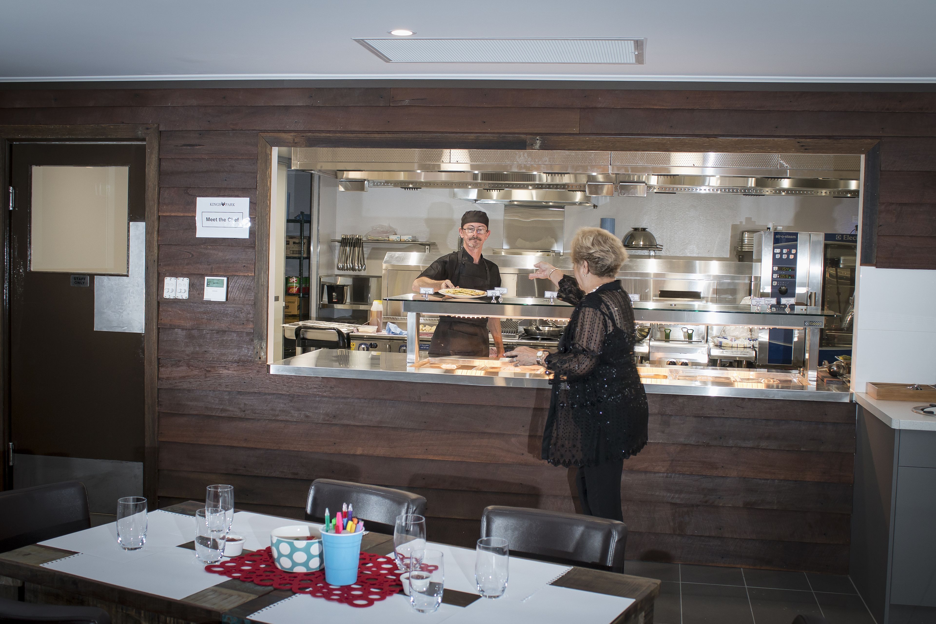 A restaurant with a wooden interior, a counter where a server is attending to a customer, and a table set with glasses and a bowl of utensils.