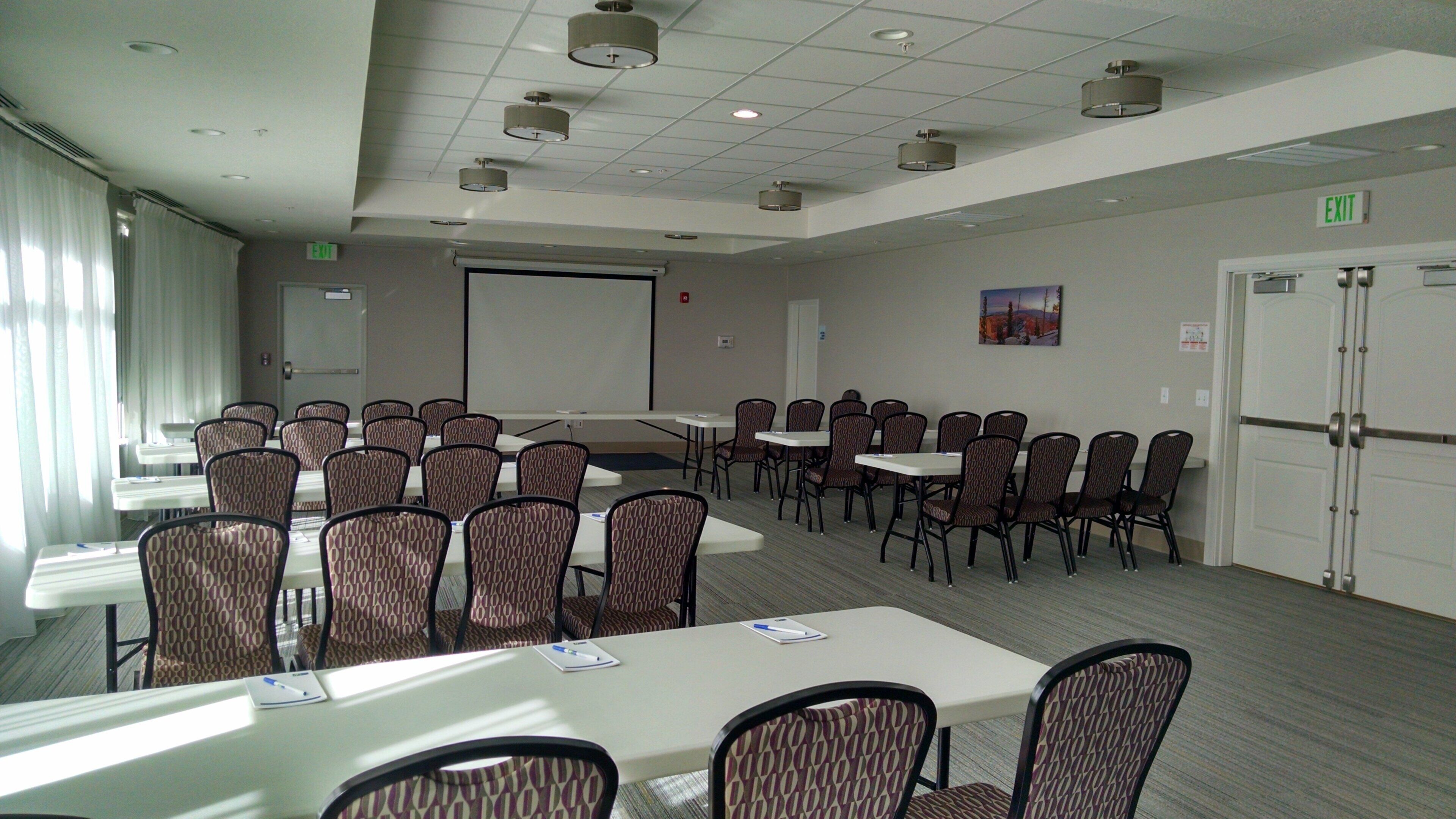A conference room with tables and chairs, a projector screen, and an exit sign.