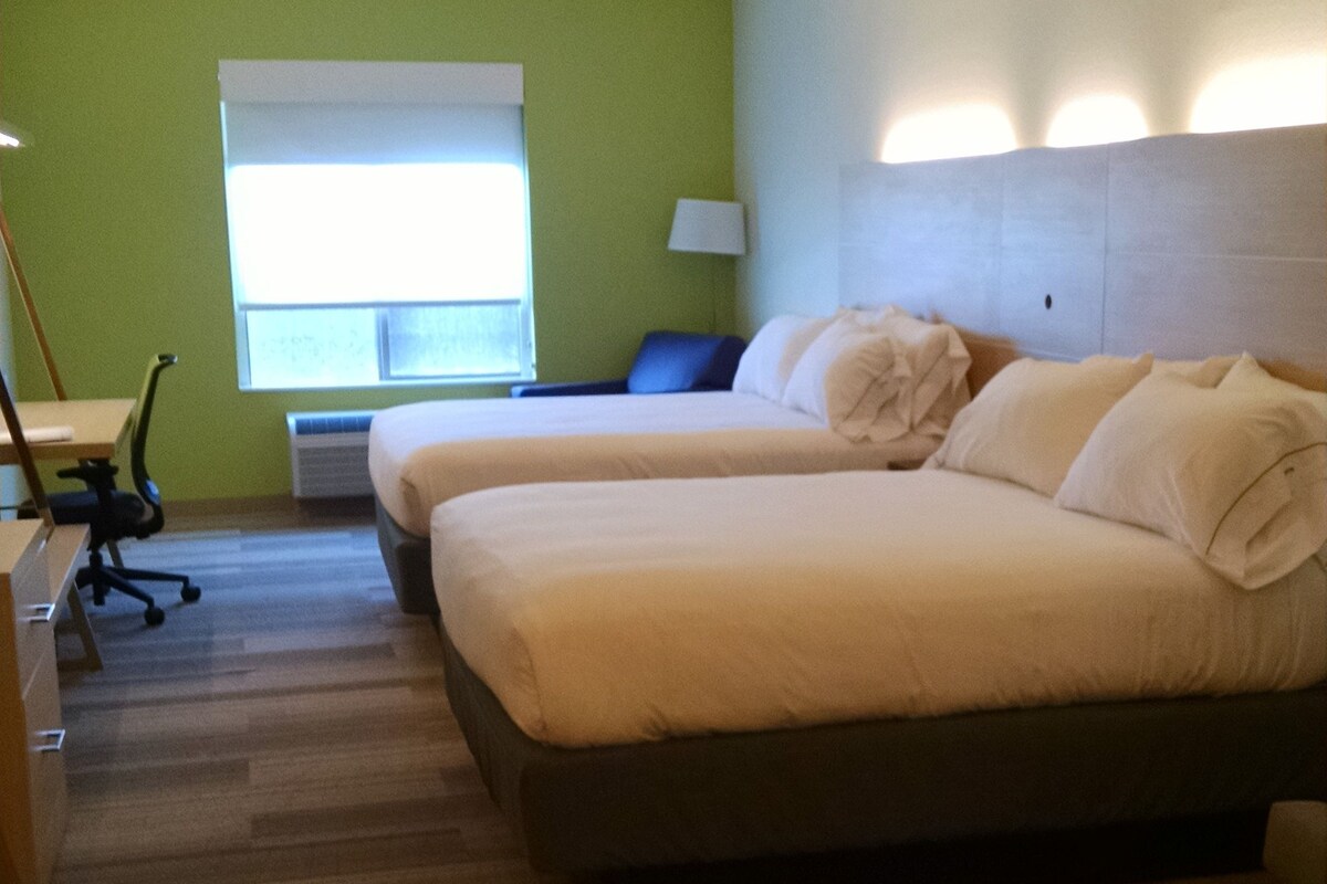 A hotel room with two beds, a desk, a chair, a lamp, and a window with blinds.