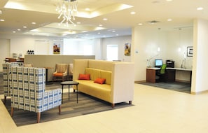 A modern hotel lobby with a yellow sofa, patterned armchairs, a small table, and a desk with a computer.