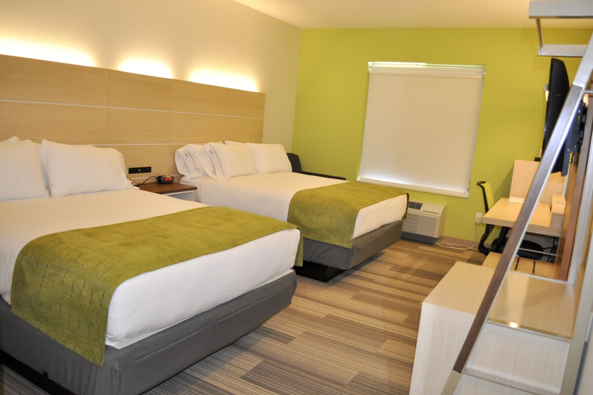 A hotel room with two beds, a desk, a chair, and a large window with a white blind.