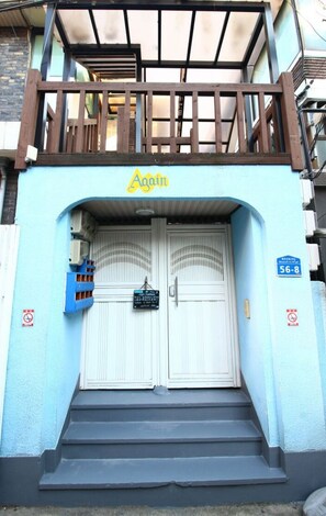 Entrada de um edifício com fachada azul, varanda de madeira e uma placa com a palavra 'Again'.