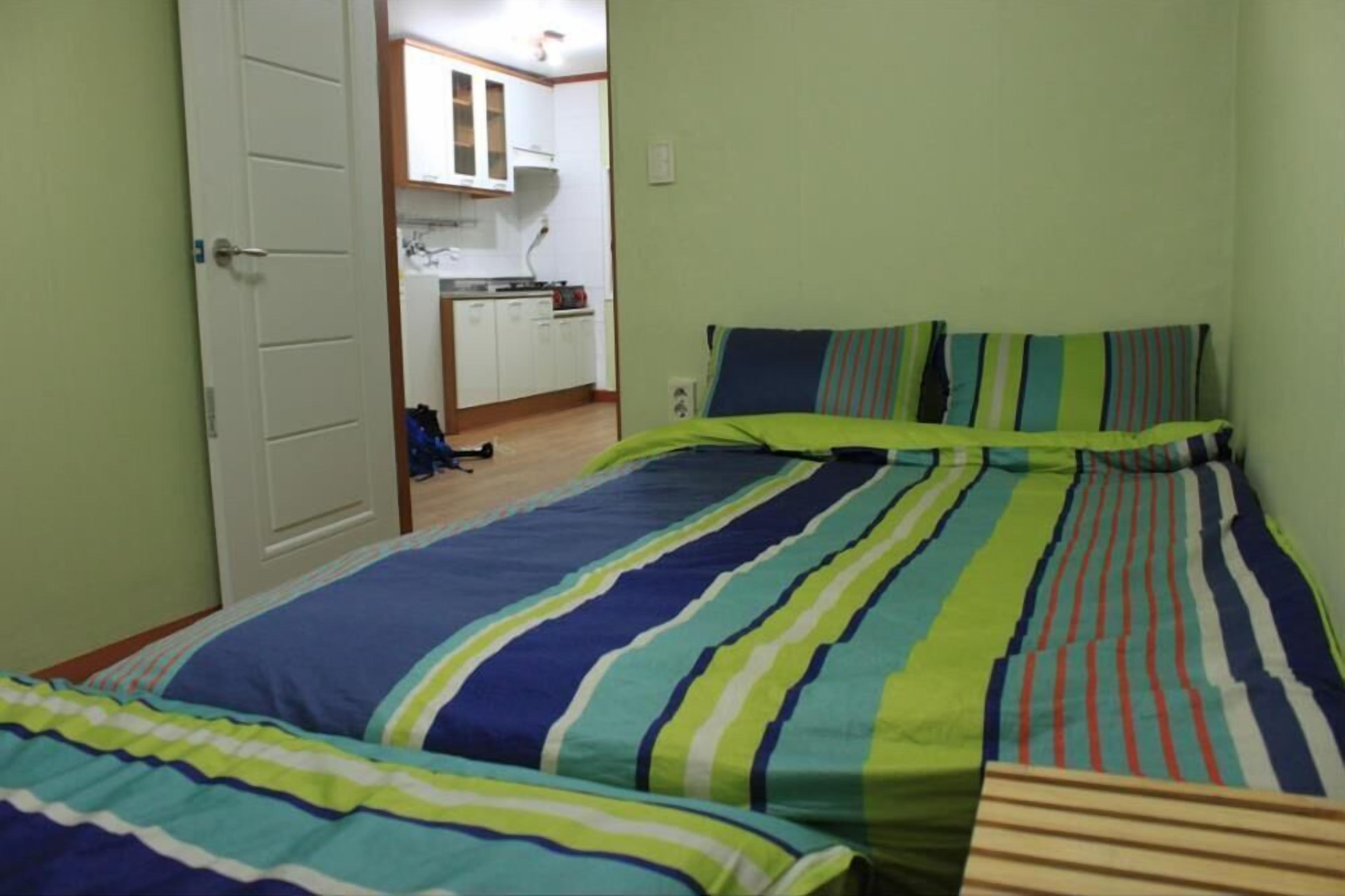 A bedroom with two beds, a visible kitchen area with cabinets and a stove, and a door leading to another room.