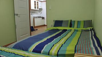 A bedroom with two beds, a visible kitchen area with cabinets and a stove, and a door leading to another room.