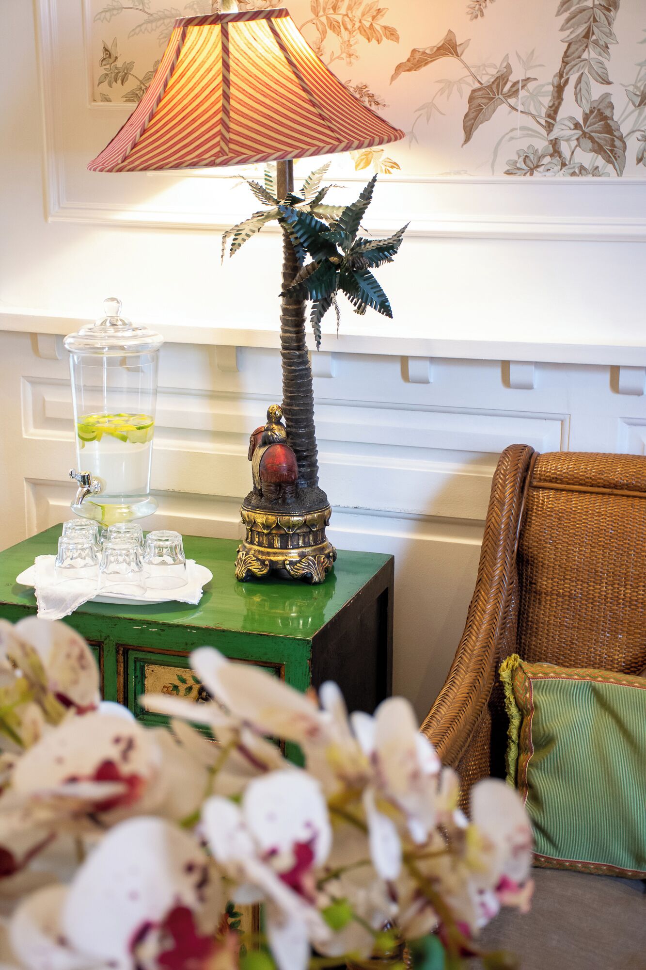 A room with a green side table holding a glass container with a drink, a decorative statue, and a wicker chair with a cushion.