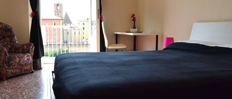 A hotel room with a bed, a chair, a small table with a vase, and a view of buildings through a window.