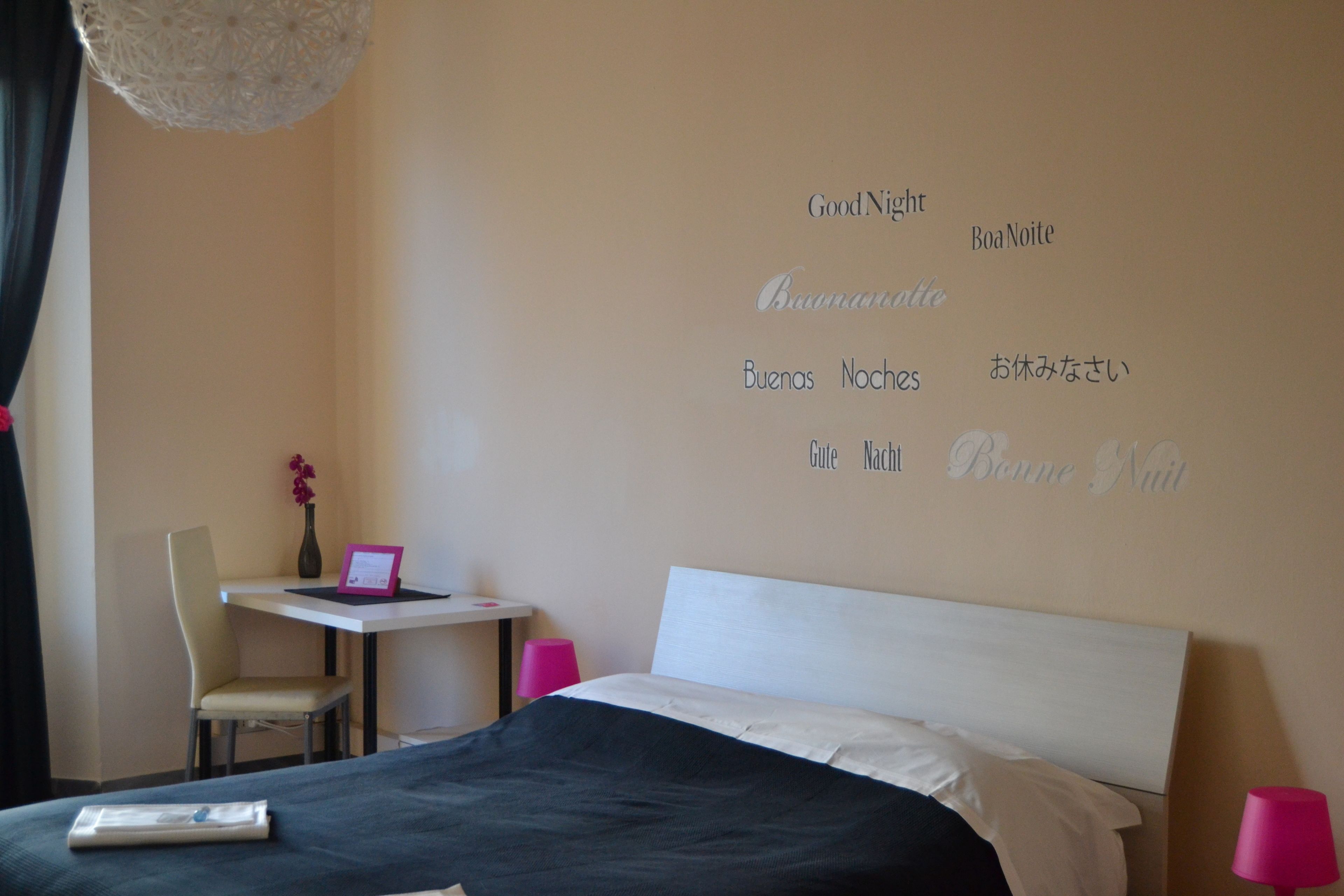 A hotel room with a bed, a desk with a laptop, a chair, and a wall with multilingual "Good Night" signs.