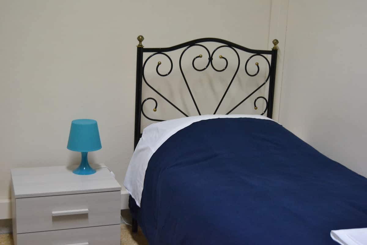 Una cama con una colcha azul oscuro y una almohada blanca, una mesita de noche con una lámpara azul, y un mueble cajonero blanco.
