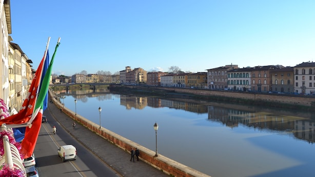 A riverside walkway with buildings on one side and a calm river in the background. - You & Me (Pisa)