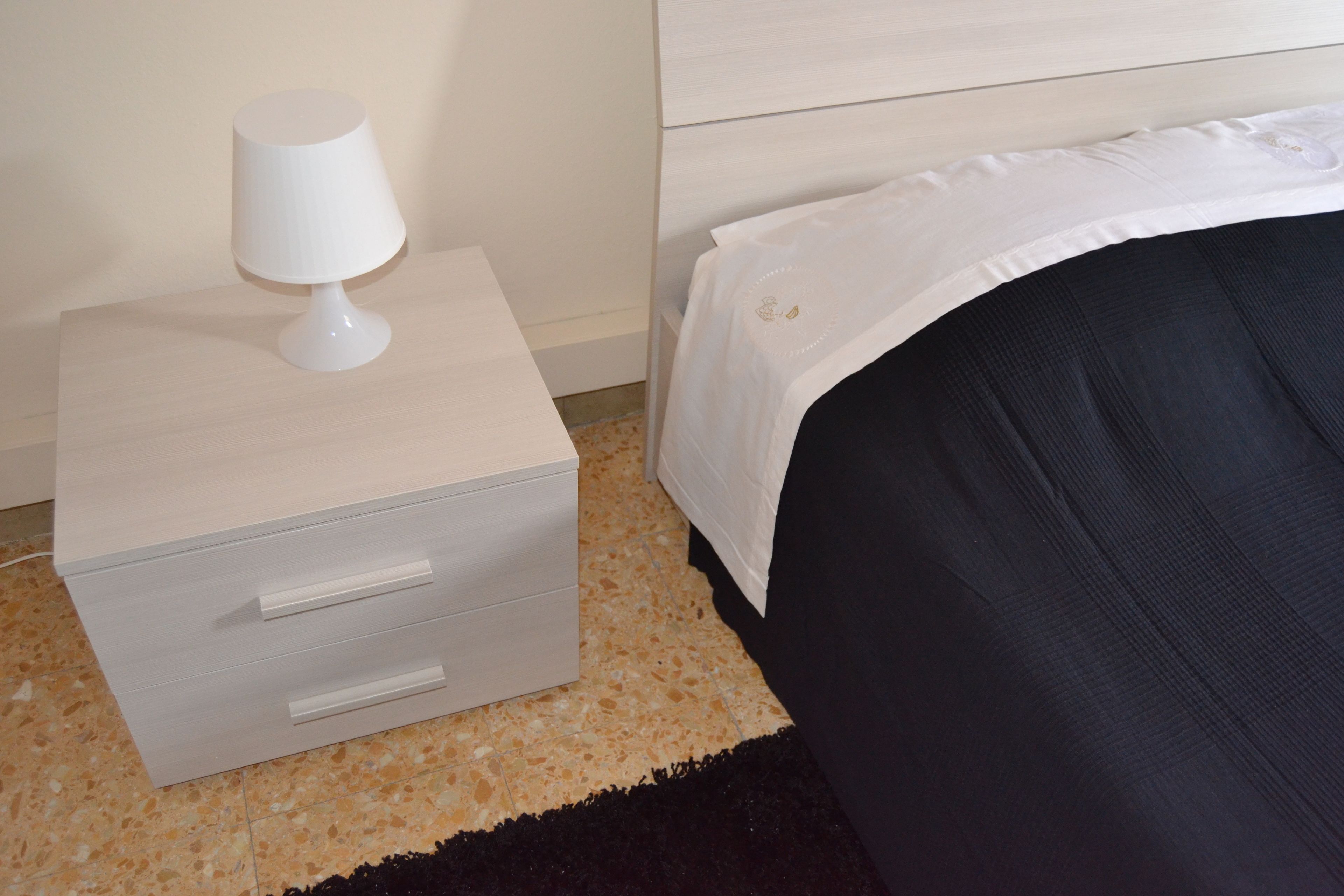 A neatly made bed with a white headboard, a small bedside table with a lamp, and a drawer unit.