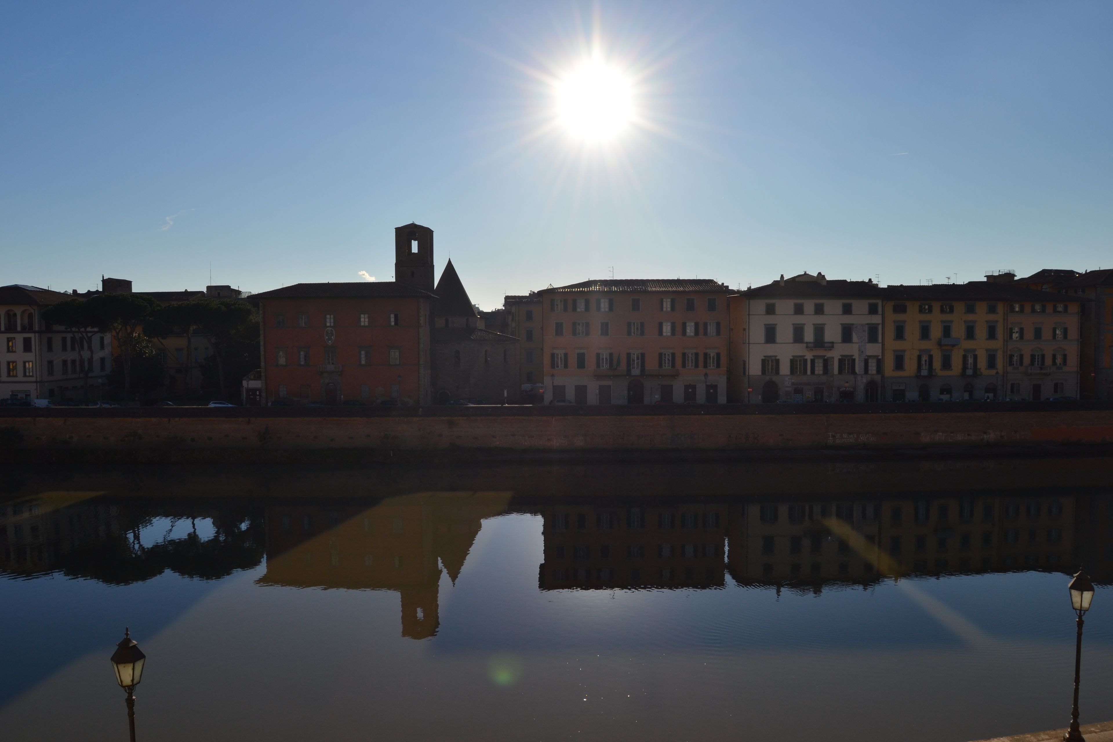 Un río tranquilo que refleja edificios, un cielo despejado y un sol brillante.