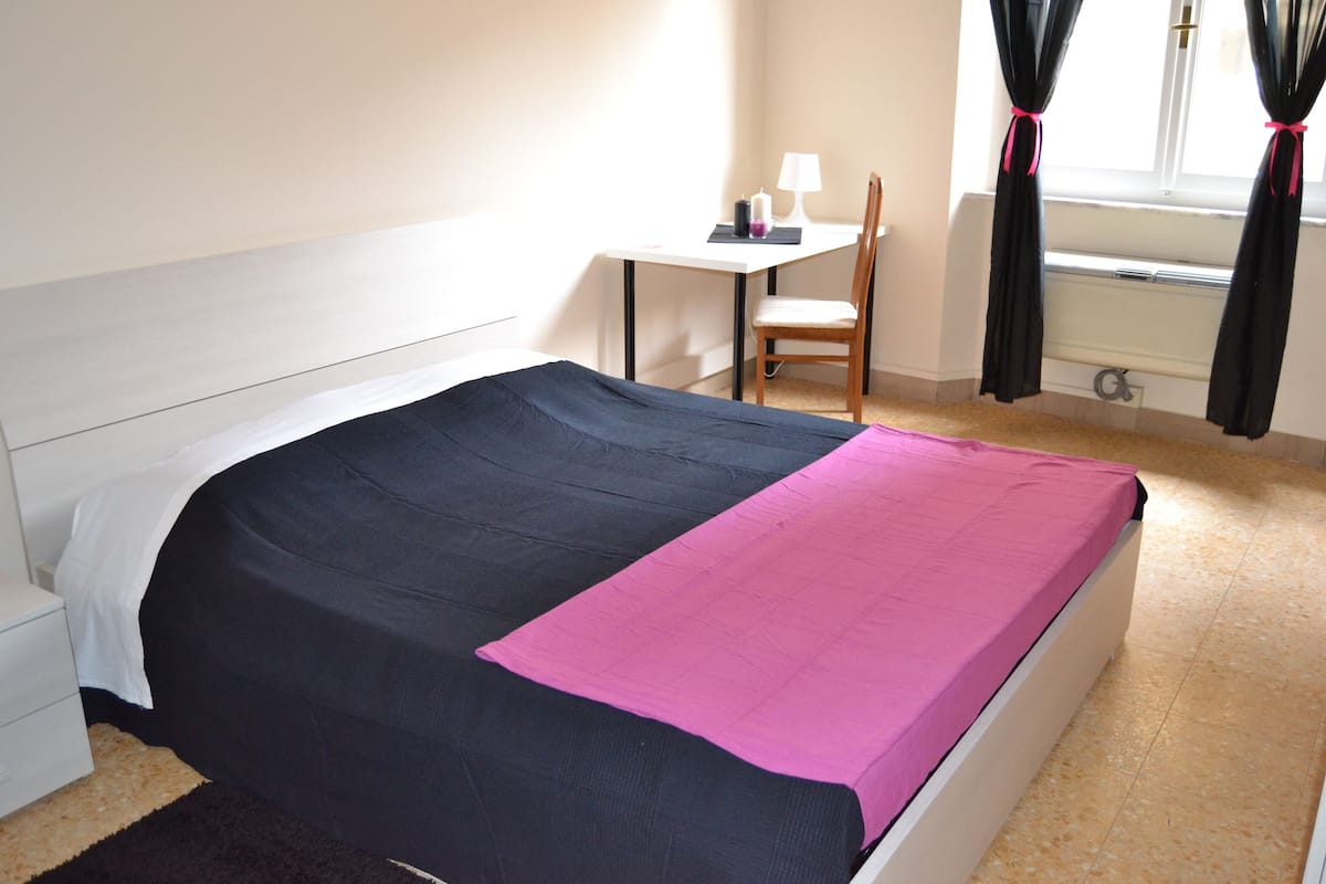 Una cama bien hecha con colcha negra y rosa, una mesita de noche con lámpara, una silla y una ventana con cortinas.