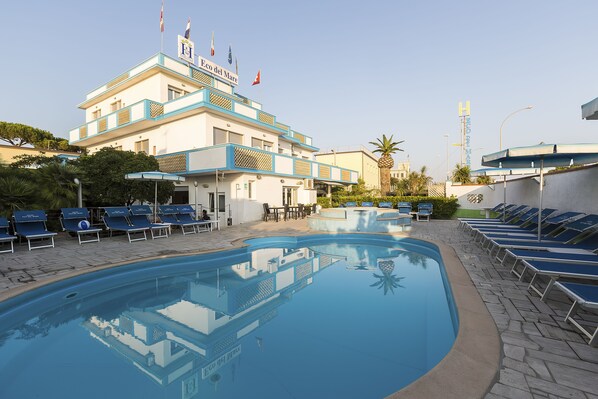 A hotel with a swimming pool, lounge chairs, and a building with flags. - Hotel Eco del Mare (Massa)