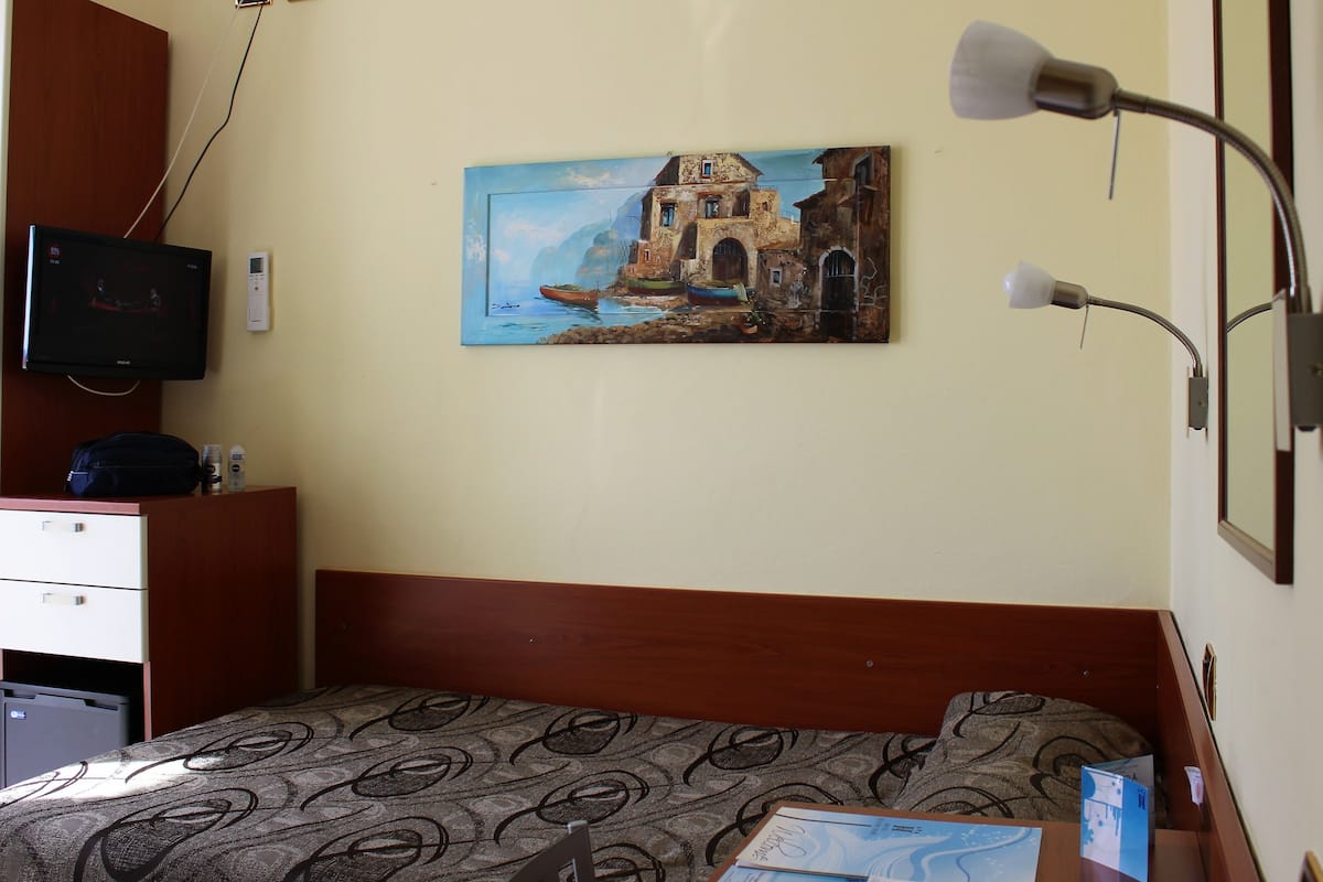 A hotel room with a bed, a desk with a computer, a painting of a coastal scene, and a lamp.