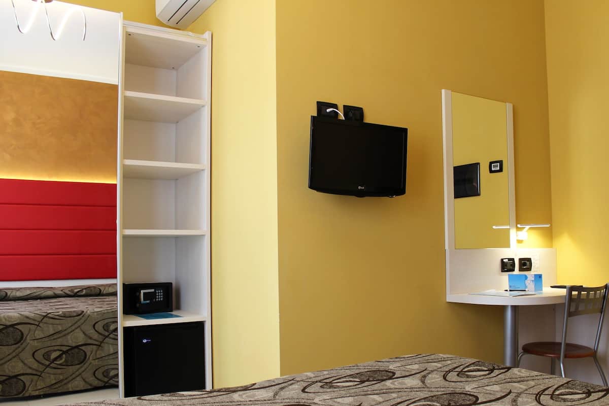 A hotel room with a wardrobe, a bed with patterned bedding, a desk with a chair, a flat-screen TV, and a mirror.