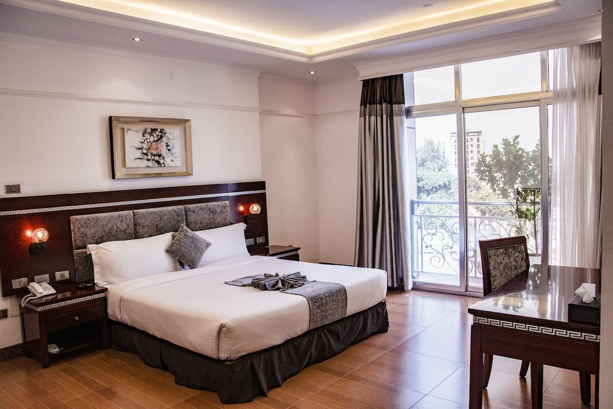 A hotel room with a large bed, a bedside table with a phone, a framed artwork on the wall, and a view of the outdoors through a large window.