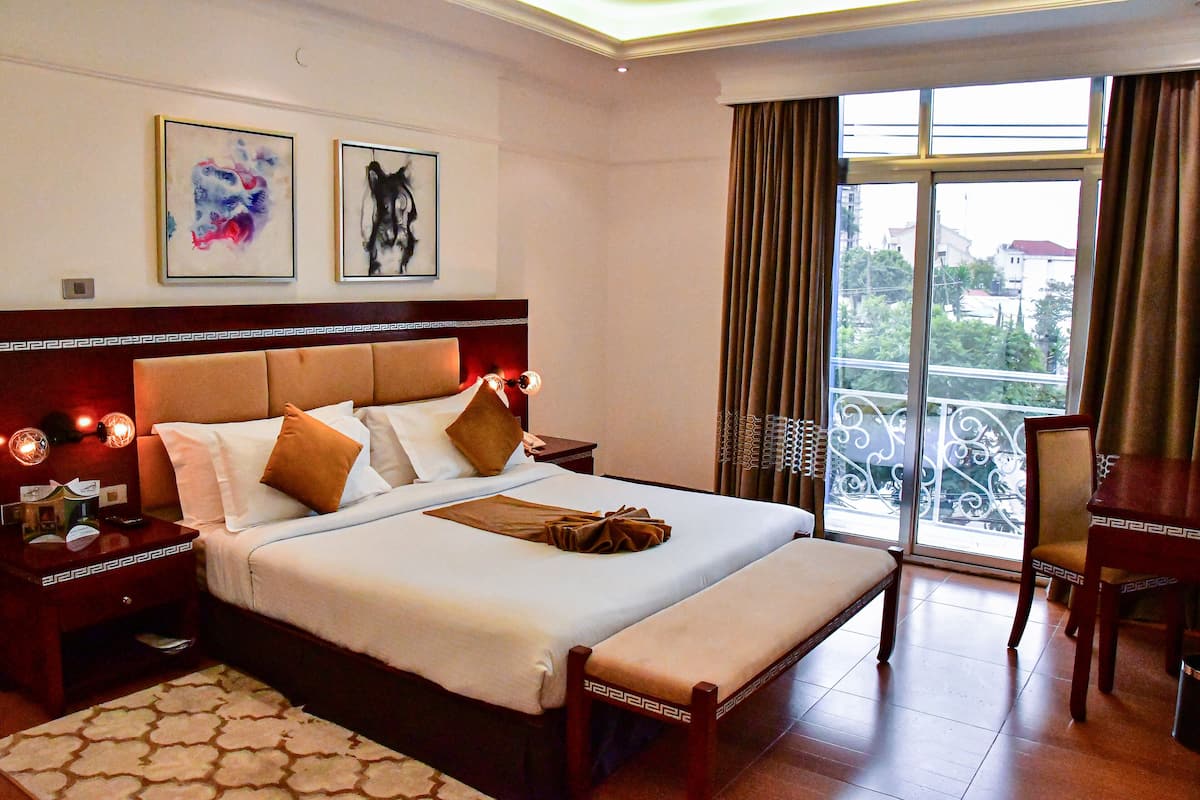 A hotel room with a large bed, a bedside table, a chair, a balcony with a railing, and two framed artworks on the wall.
