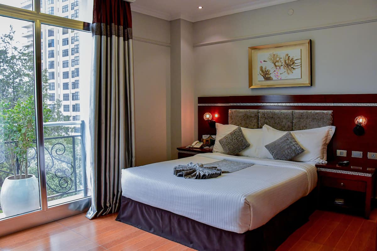 A hotel room with a large bed, a nightstand, a wall-mounted lamp, a framed artwork, and a view of a building through a window.