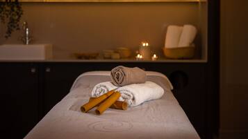 A massage table with towels and candles, a sink, and a shelf with candles and toiletries.