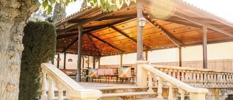 A covered outdoor seating area with steps leading up to it, surrounded by stone walls and a tree.