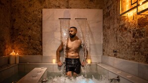 A man in a bathtub with water flowing over him, candles on the side, and a stone wall in the background. - RVHotels Broquetas Spa Termal (Caldes de Montbui)