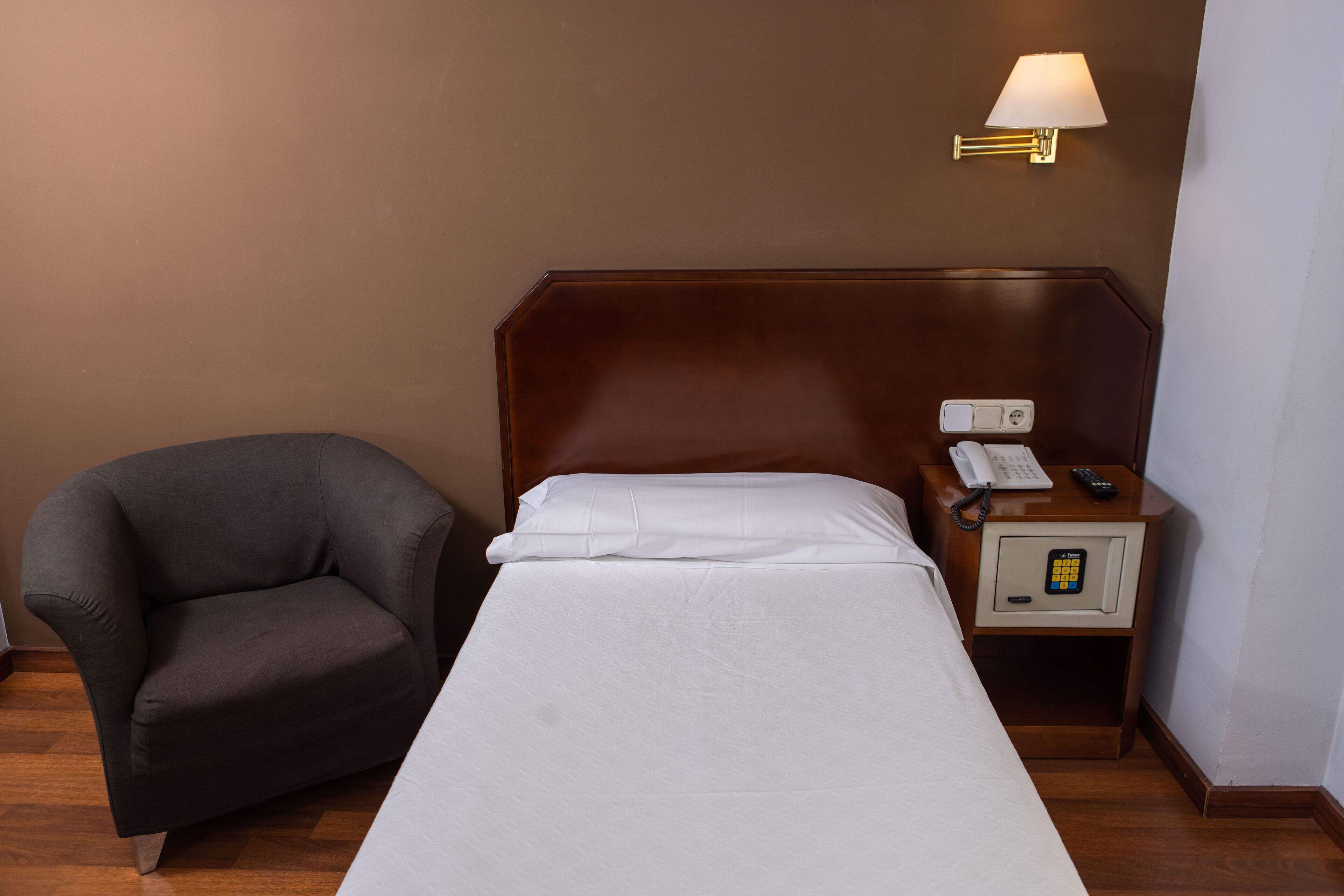 Una habitación de hotel con una cama individual, una silla, una mesita de noche con lámpara y un teléfono.