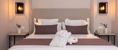 A neatly made bed with a brown headboard, white pillows, and a white bathrobe on the bed. There are two bedside tables with lamps and a small plant on each.
