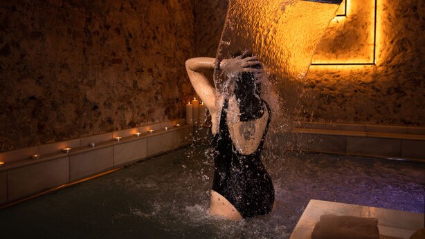 A person in a black swimsuit in a water feature with a stone wall background and candles along the edge. - RVHotels Broquetas Spa Termal (Caldes de Montbui)
