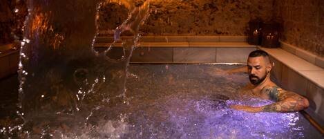 A man in a hot tub with water jets, in a room with stone walls and a shelf with candles.