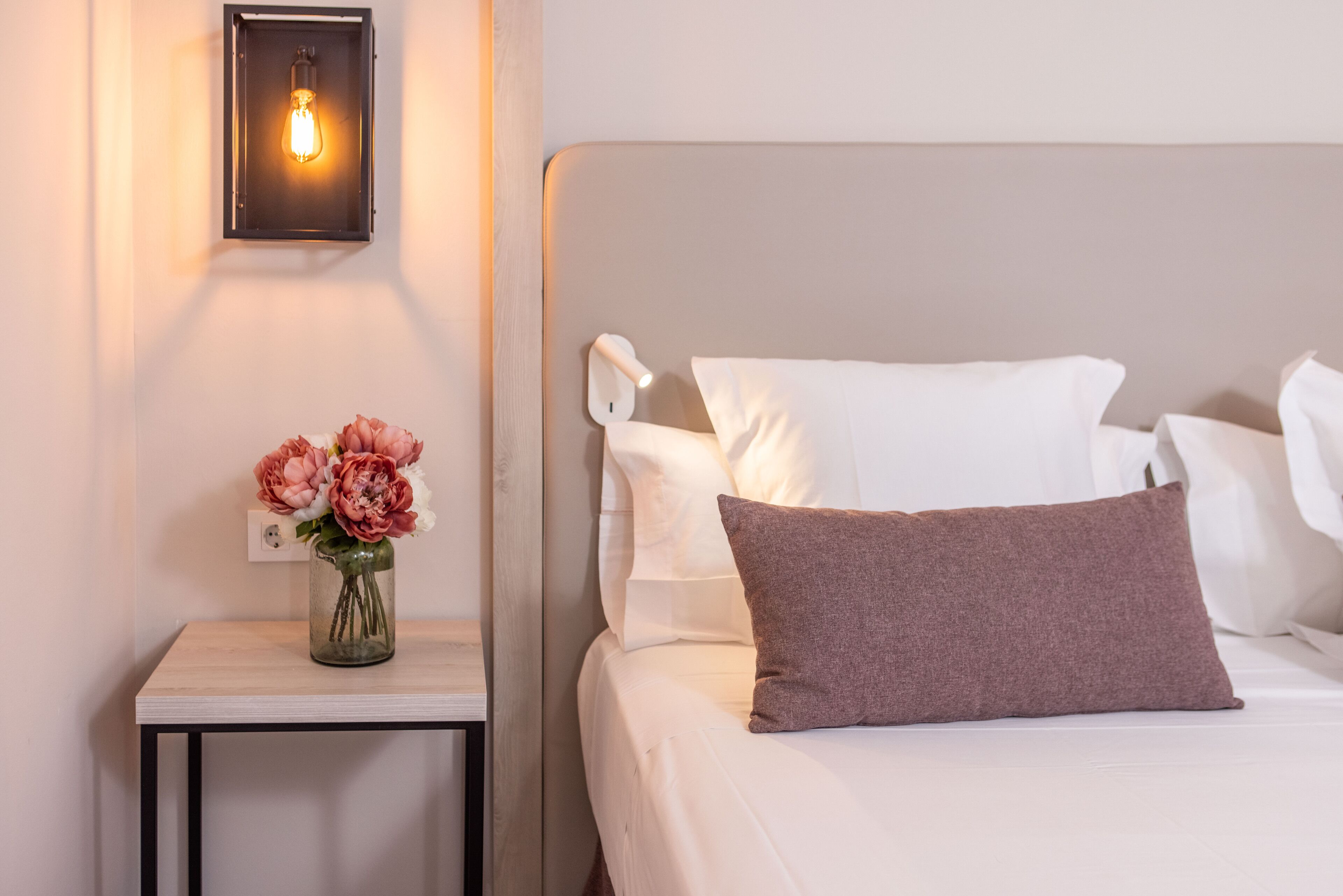 Een opgemaakt bed met witte en bruine kussens, een vaas met bloemen op een nachtkastje, en een wandlamp.