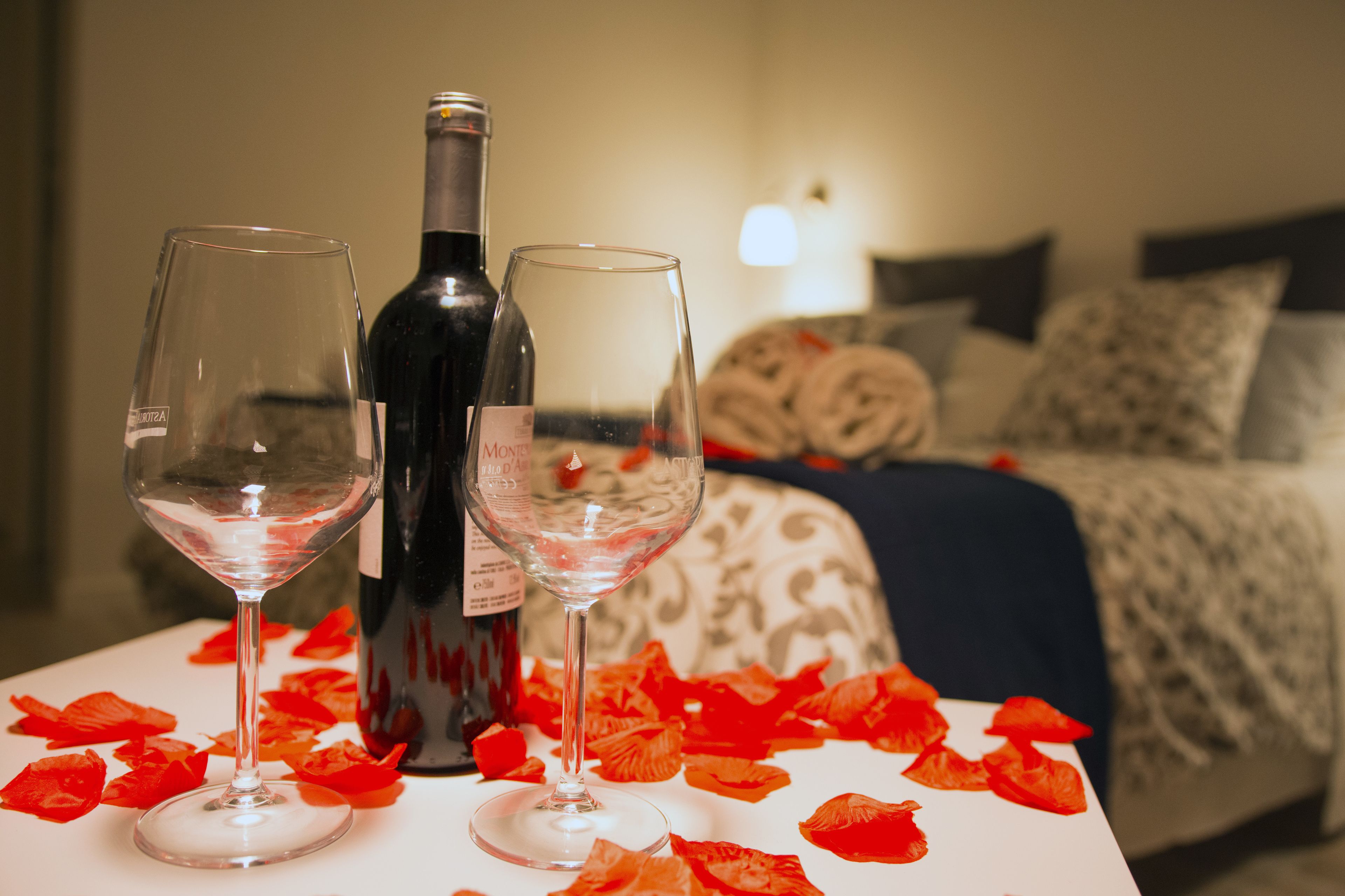 A bedroom with a bed, pillows, and a bedside table with a wine bottle and two glasses.