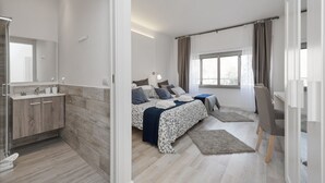 A modern bedroom with a wooden floor, a bed with a patterned duvet, a bedside table, a chair, and a window with curtains. - De Guestibus (Rome)