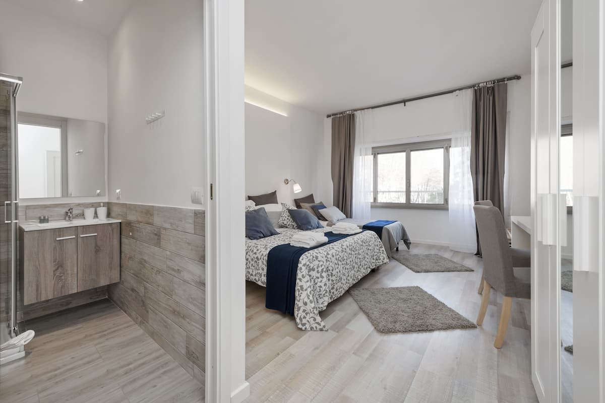 A modern bedroom with a wooden floor, a bed with a patterned duvet, a bedside table, a chair, and a window with curtains.